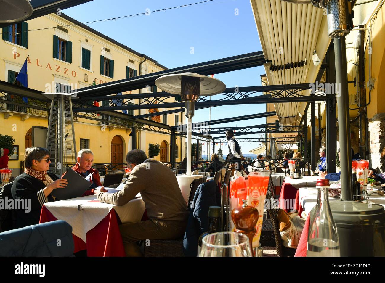 People and tourists sitting in an outdoor restaurant in the old town on the shore of Lake Garda in a sunny day, Lazise, Verona, Veneto, Italy Stock Photo