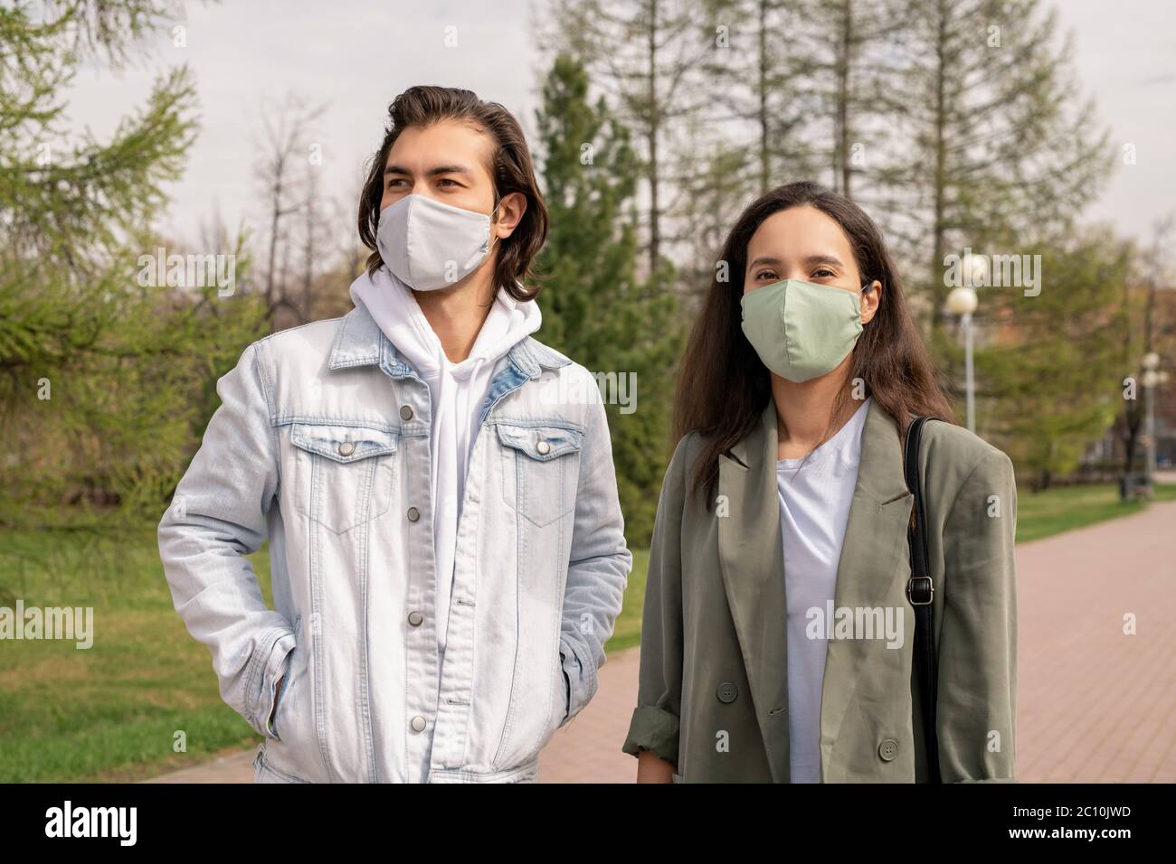 Young couple in facial masks walking together in city park during coronavirus epidemic Stock Photo