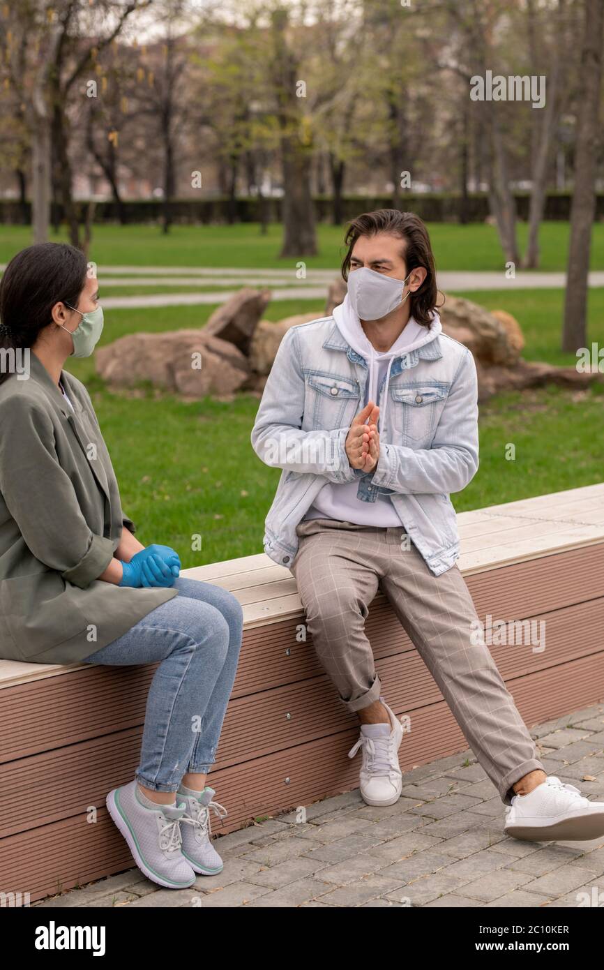 Young friends sitting on bench in city park and chatting while spending free time outdoors during pandemic Stock Photo