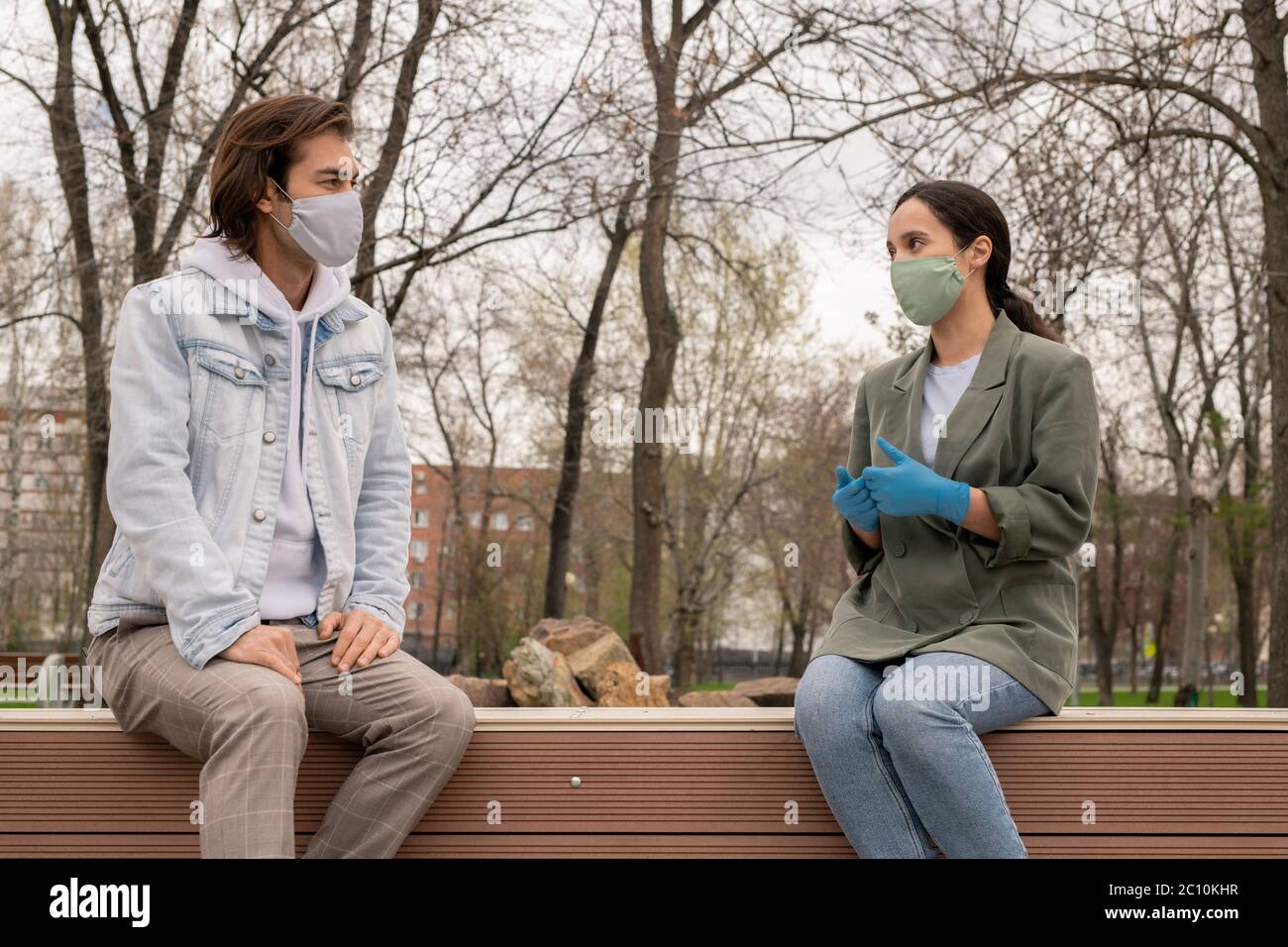 Young couple sitting on bench in park and keeping distance while talking to each other during coronavirus period Stock Photo