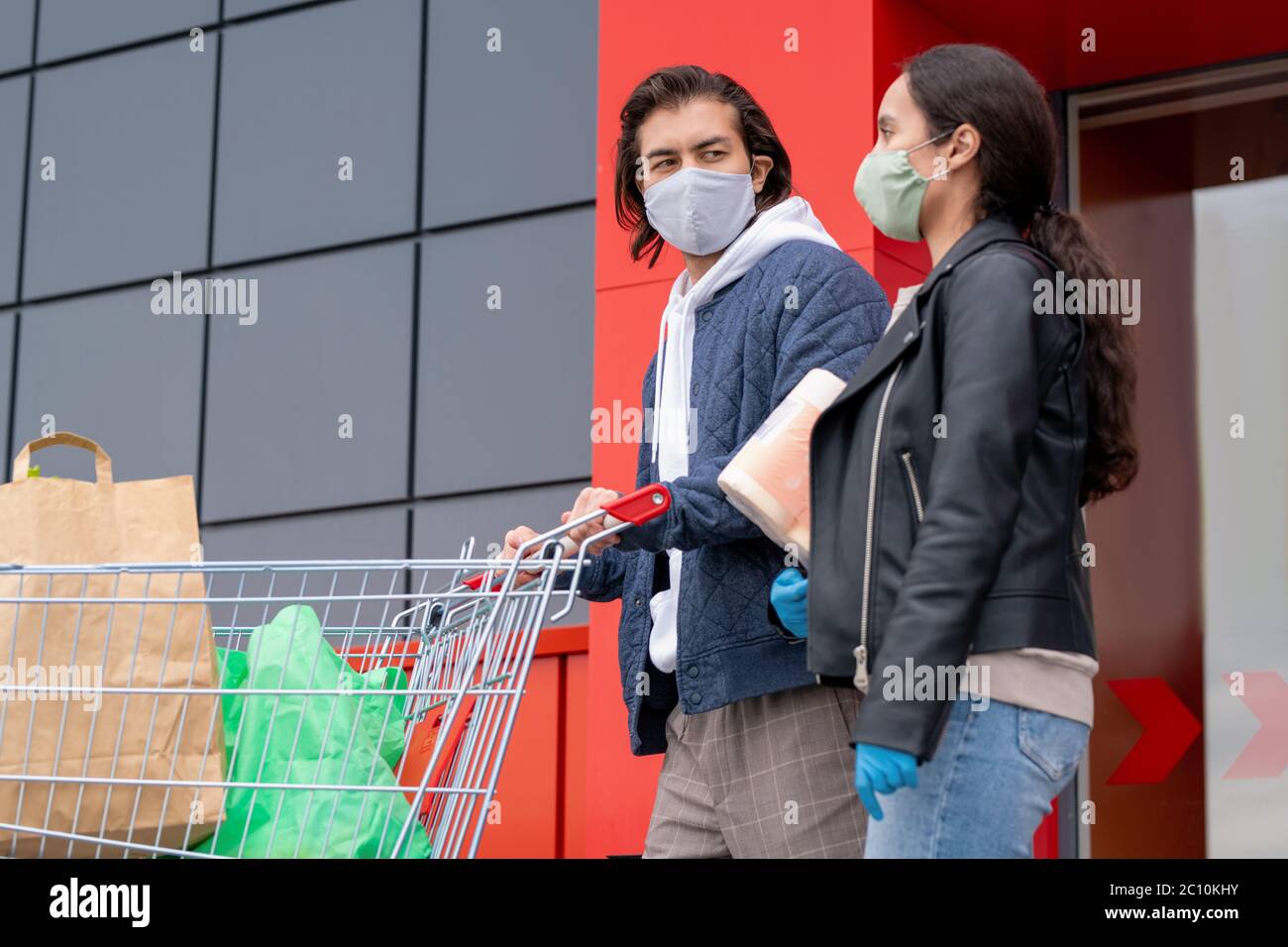 Young couple in jackets and cloth mask leaving supermarket together after shopping, man pushing cart Stock Photo