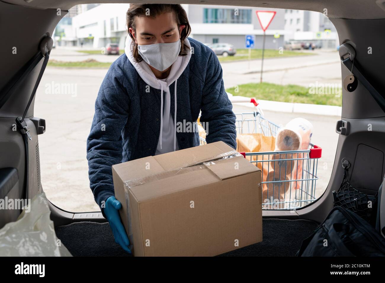 Middle-aged man in cloth mask and jacket standing at car and loading box into trunk after visiting store Stock Photo