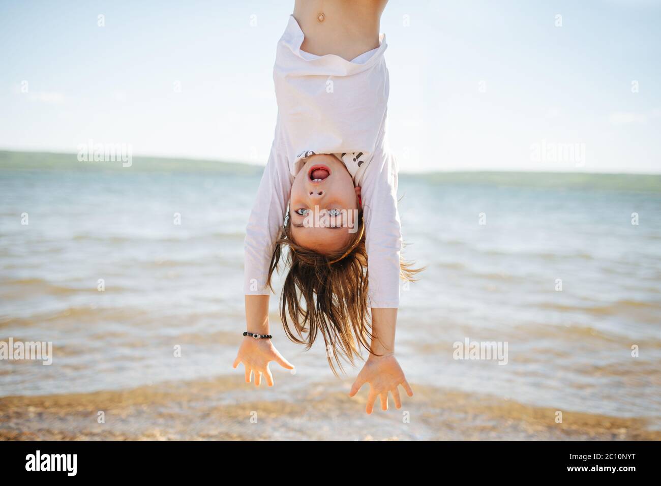 Little hiperemotional school age girl hanging upside down. On a lake shoreside. Stock Photo