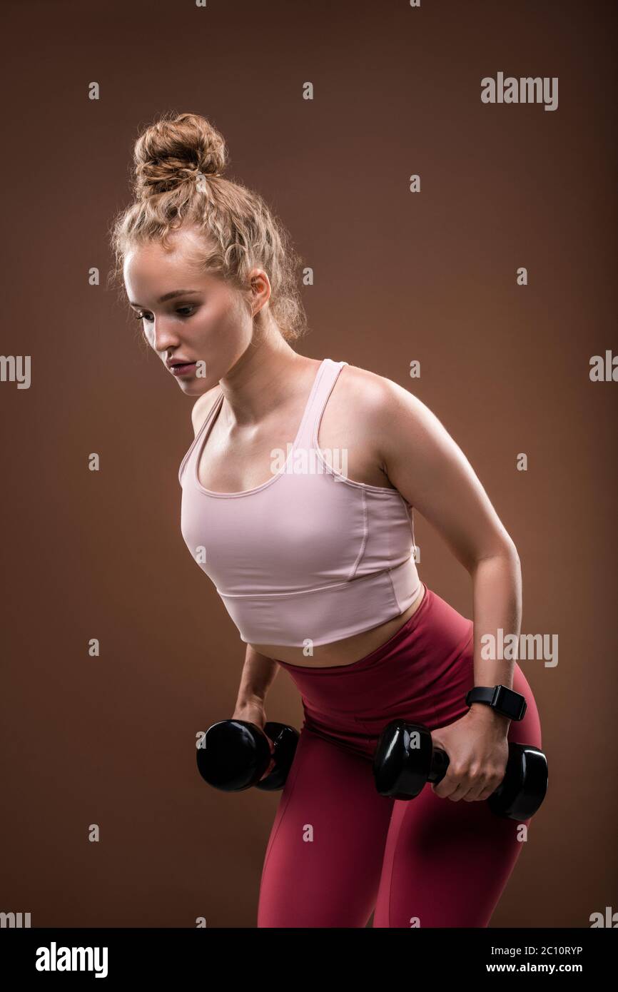 Young blond female in tracksuit bending slightly forwards while doing exercise for arms with dumbbells during workout in isolation Stock Photo