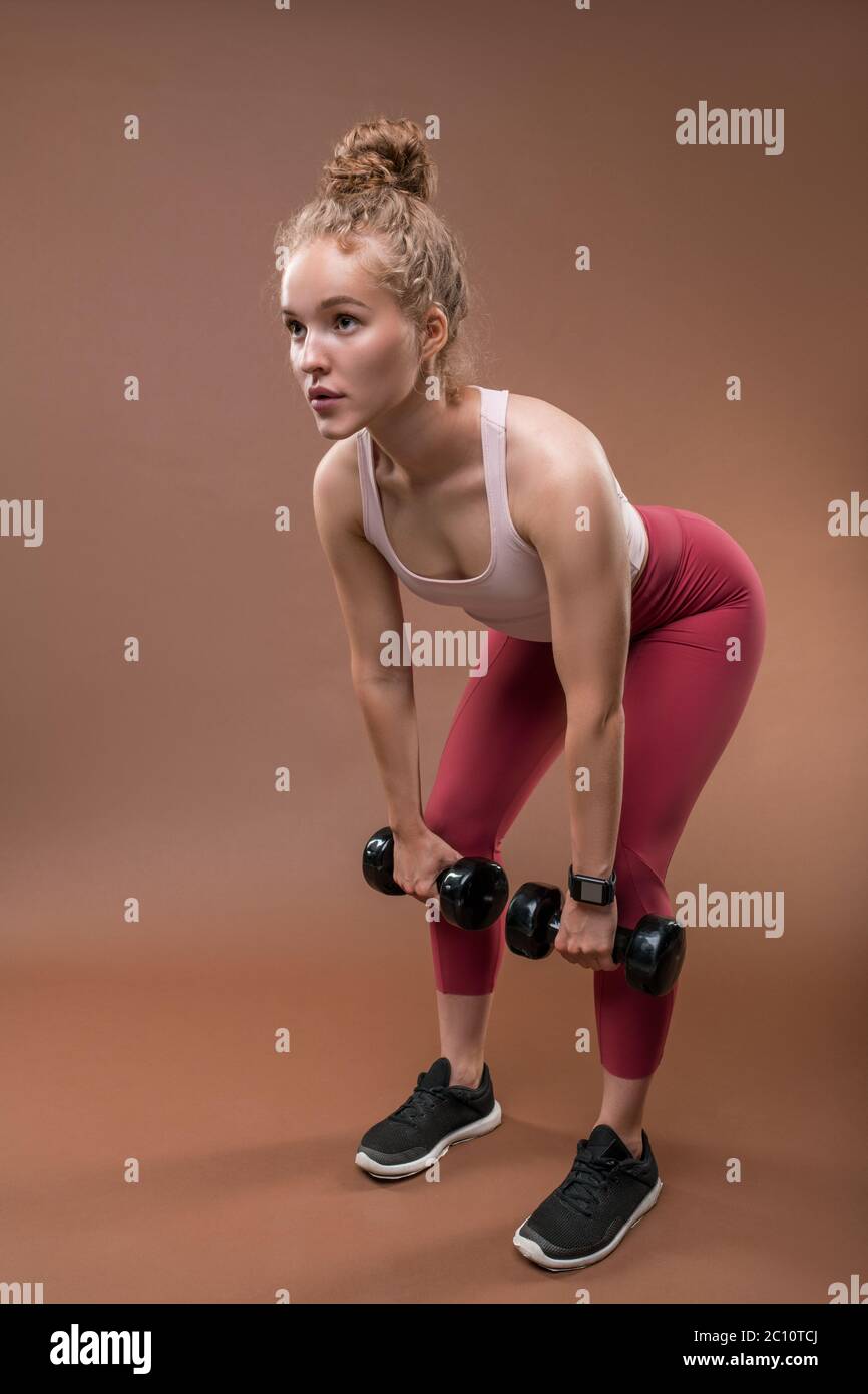 Young blond female athlete in activewear bending slightly forwards with her legs bent in knees while lifting dumbbells during training Stock Photo