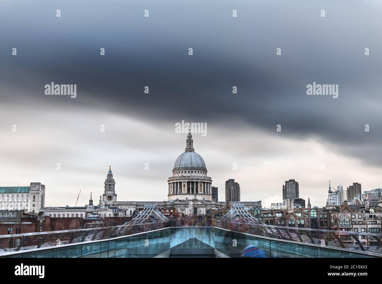 Crowds of locals and tourists cross the river Thames in London over a footbridge, with the dome of St. Paul’s Cathedral under menacing rain clouds. Lo Stock Photo