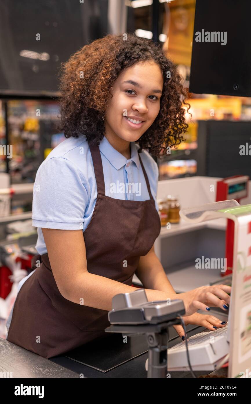 Happy young mixed-race cashier with dark wavy hair looking at you while standing by workplace and pressing button of cashbox Stock Photo