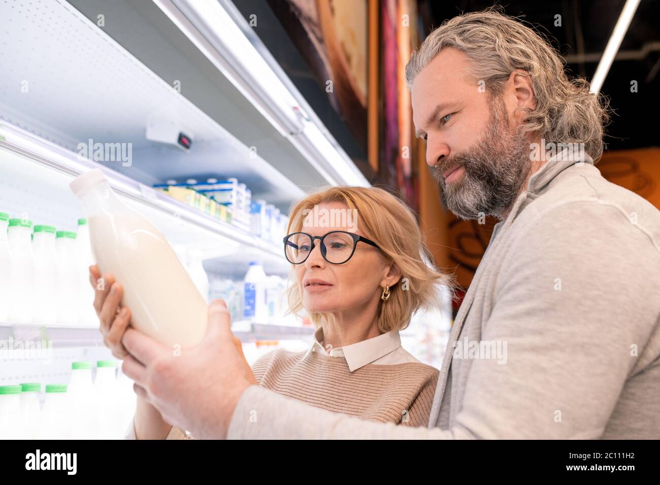 Aged serious bearded man and his blond wife choosing milk on display with dairy products while standing by shelves with bottles Stock Photo
