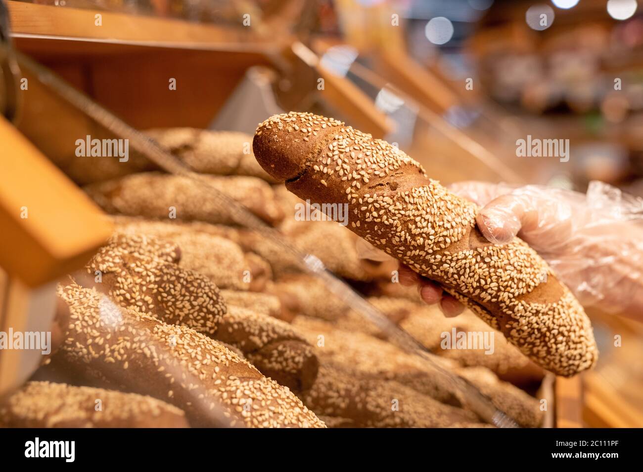 Gloved hand of mature female buyer holding fresh bread sprinkled with sesame seeds while standing by display in supermarket Stock Photo