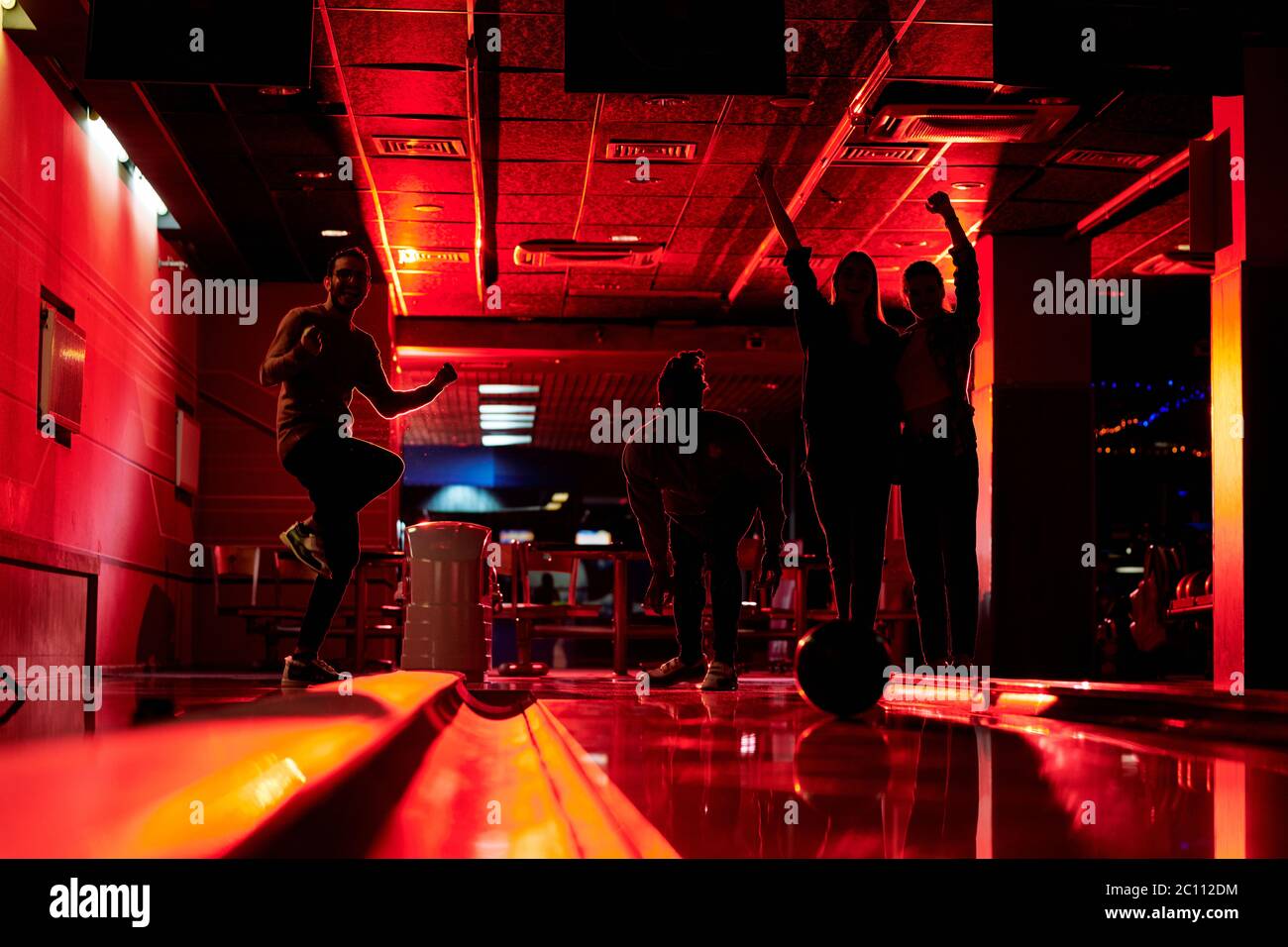 Silhouettes of young friends expressing gladness while one of them throwing ball on bowling track in dark room of entertainment club Stock Photo