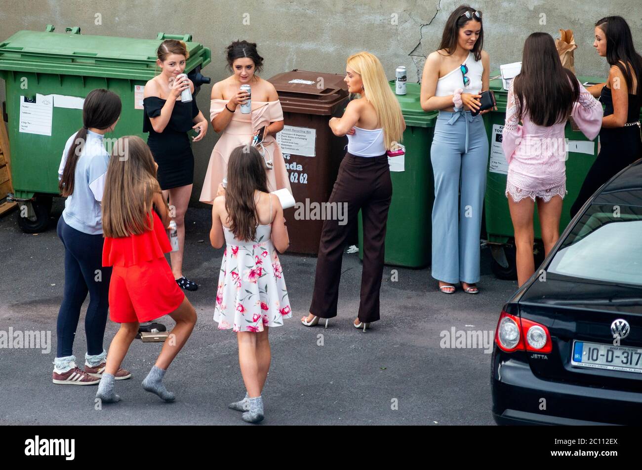 Young women and teenagers in smart prom dresses drinking alcohol beer illegally in back garden public car park parking lot before going to their graduation party as a social problem issue. Stock Photo