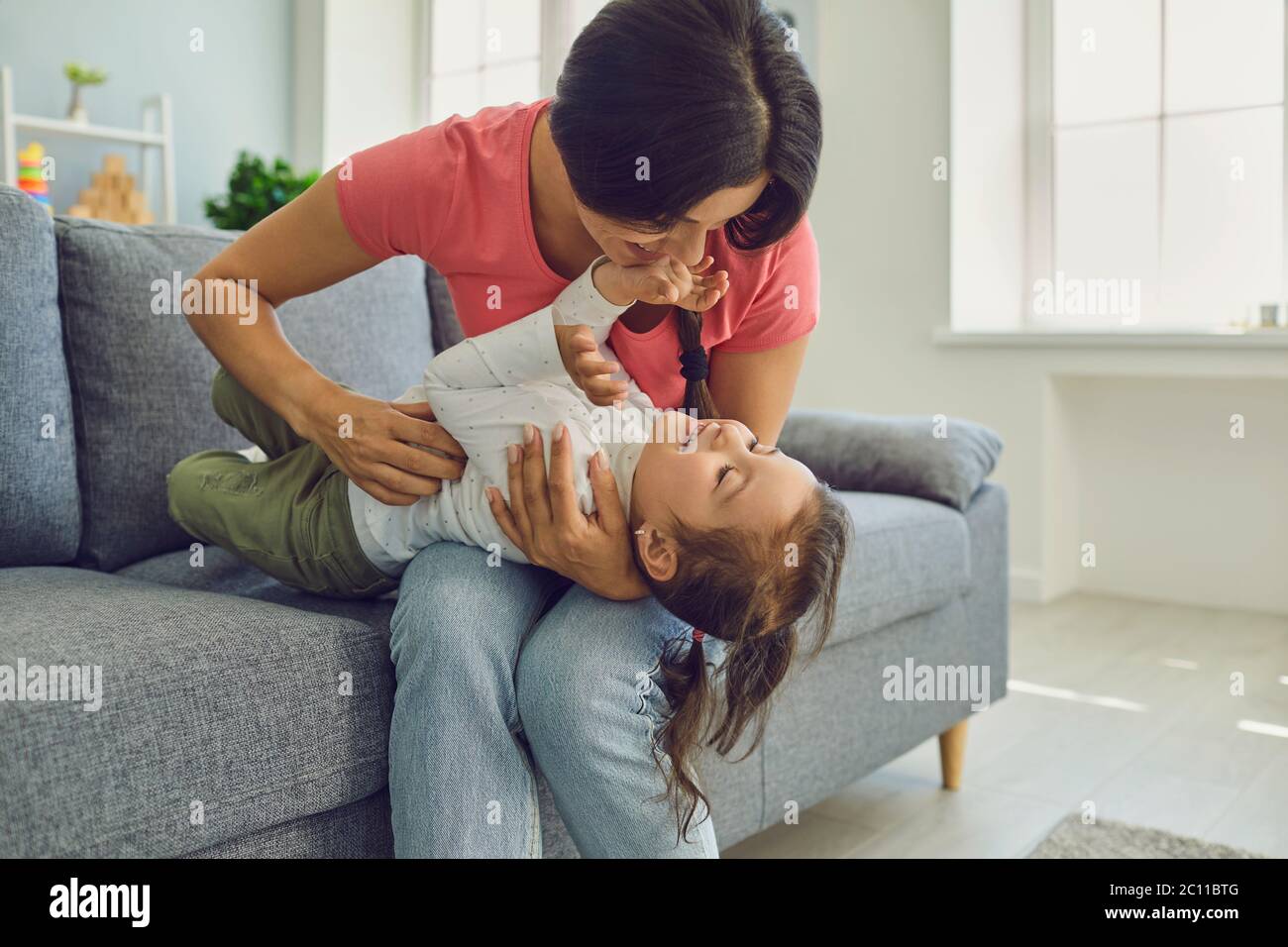 Happy mother and daughter hugging and playing on living room sofa. Family epidemic home isolation. Stock Photo