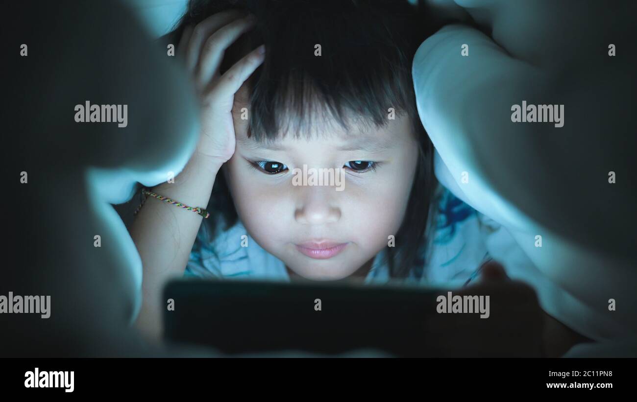 Eye close up Little Girl are watching video in tablet on bed at night time light flashes reflected from the screen,children using games with addiction Stock Photo