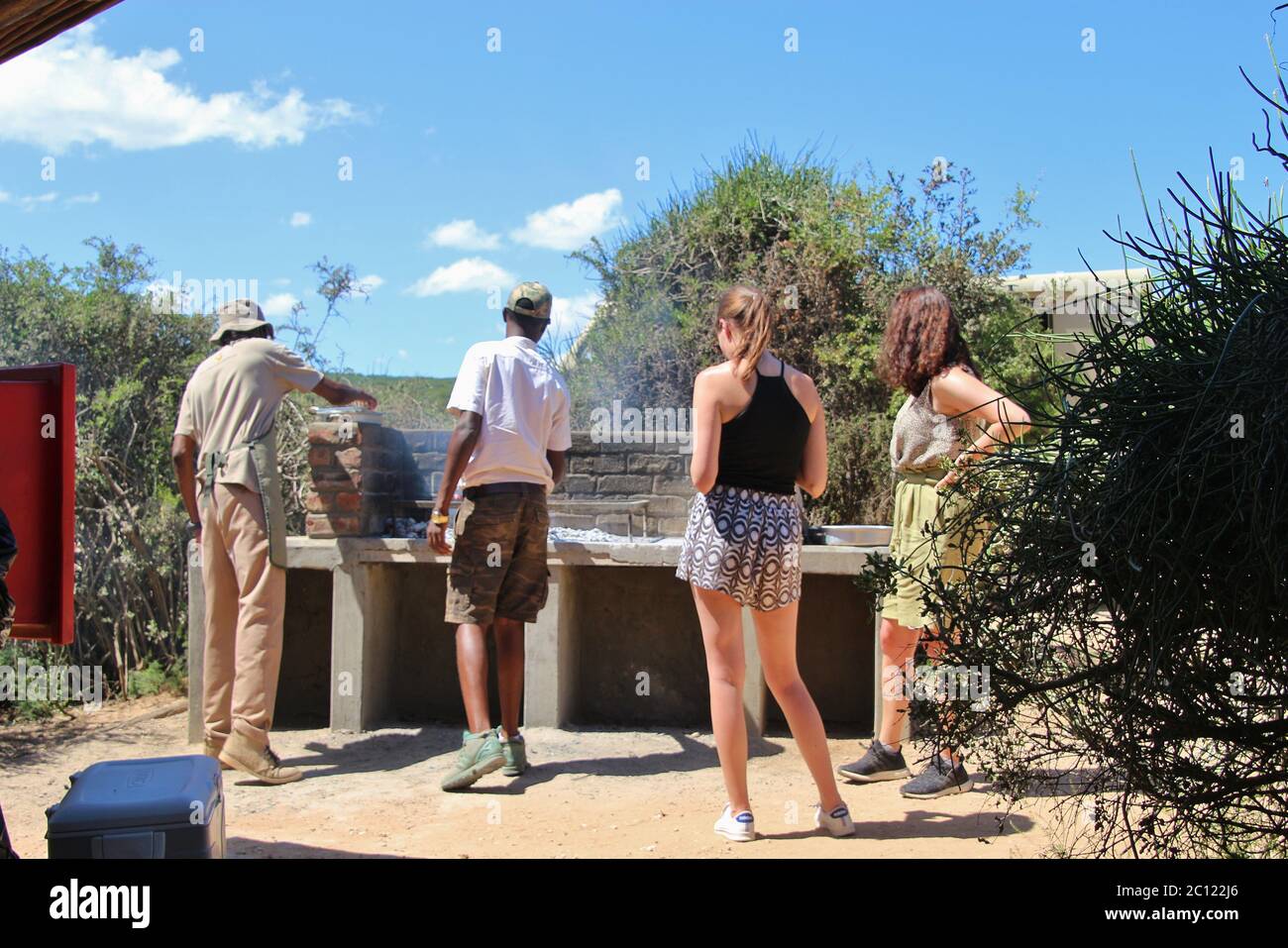 Two safari guides grill meat for the safari participants at a picnic site in Addo Elephant Park. Two watching tourists besides the grill. Africa. Stock Photo