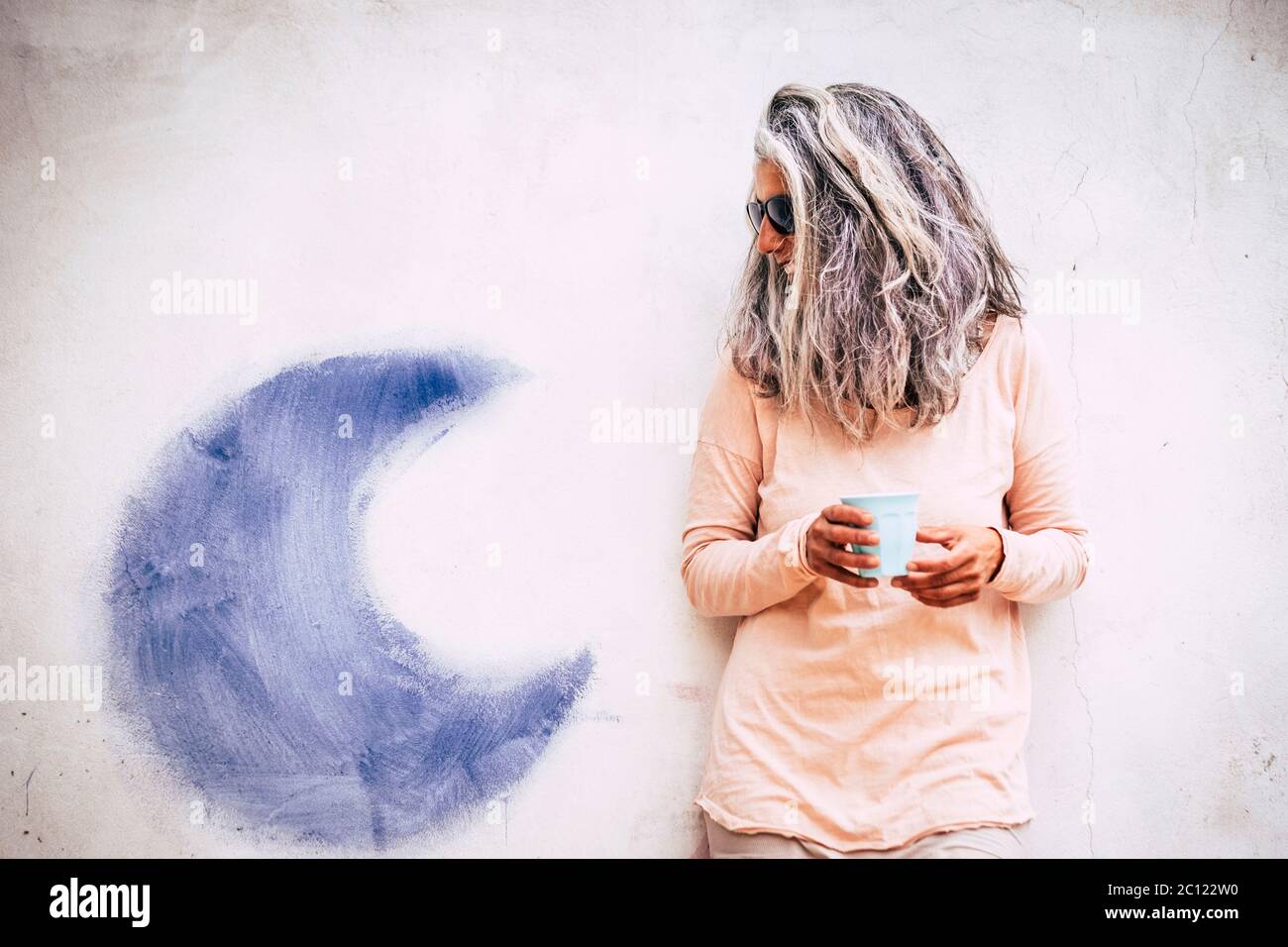 Portrait of adult senior alternative caucasian people woman with long grey and white hair and cup of tea .- white wall wih blue moon in background Stock Photo