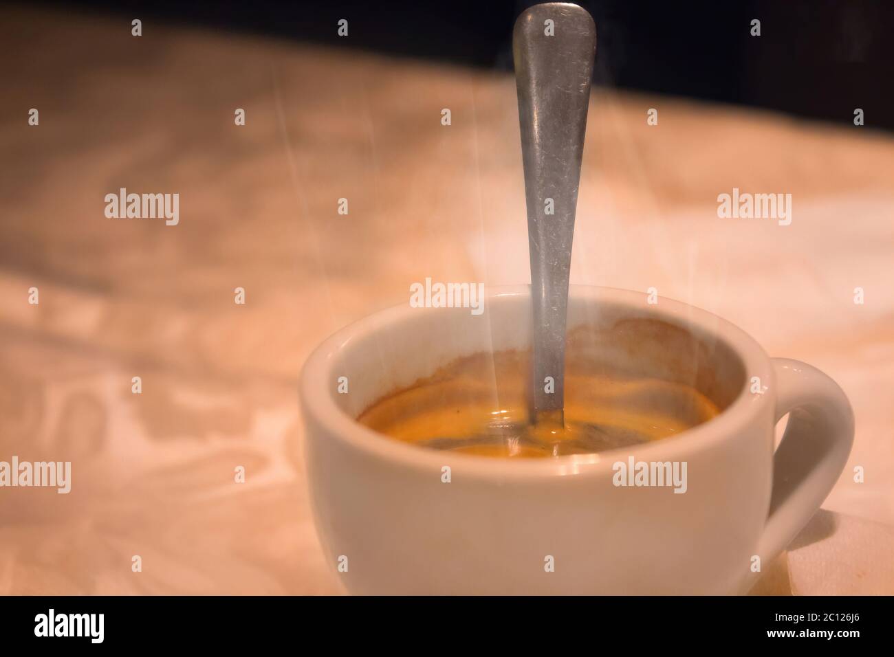 Smoke emitted from a cup with hot coffee and teaspoon in it. It placed on a tablecloth on a table. Close up with selective focus. Stock Photo