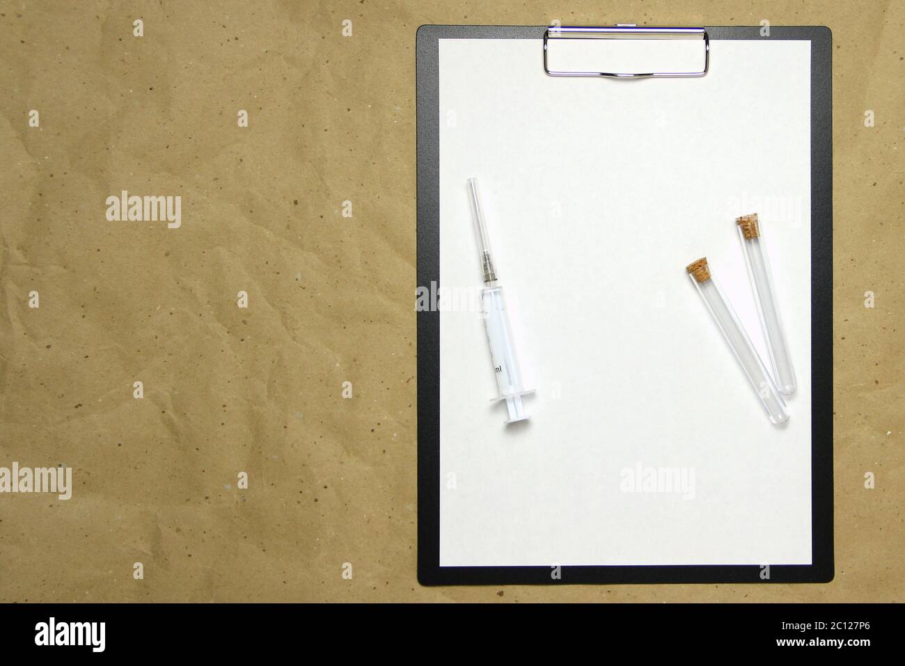 The tablet with a clip for the clip of paper with a white sheet a4 lies against the background of craft brown crumpled paper. On top is a medical syringe and two transparent empty tubes with stoppers. Place for text and layout for design. Stock Photo