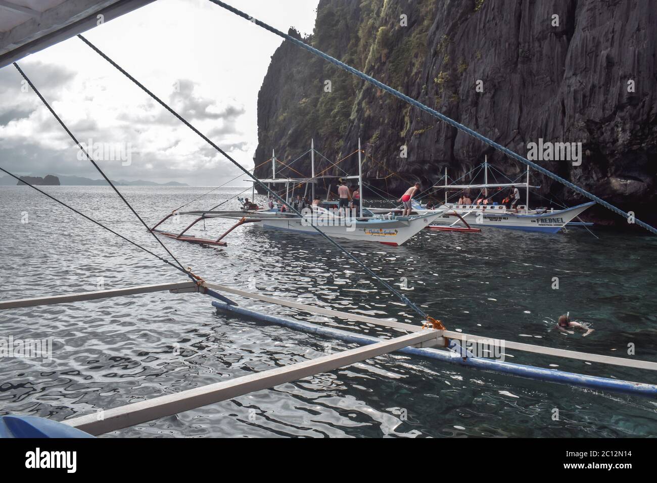 Tourists on traditional Philippino fisherman boats crossing the waters near grey and green limestone cliffs on a cloudy day of El Nido at Palawan Isla Stock Photo