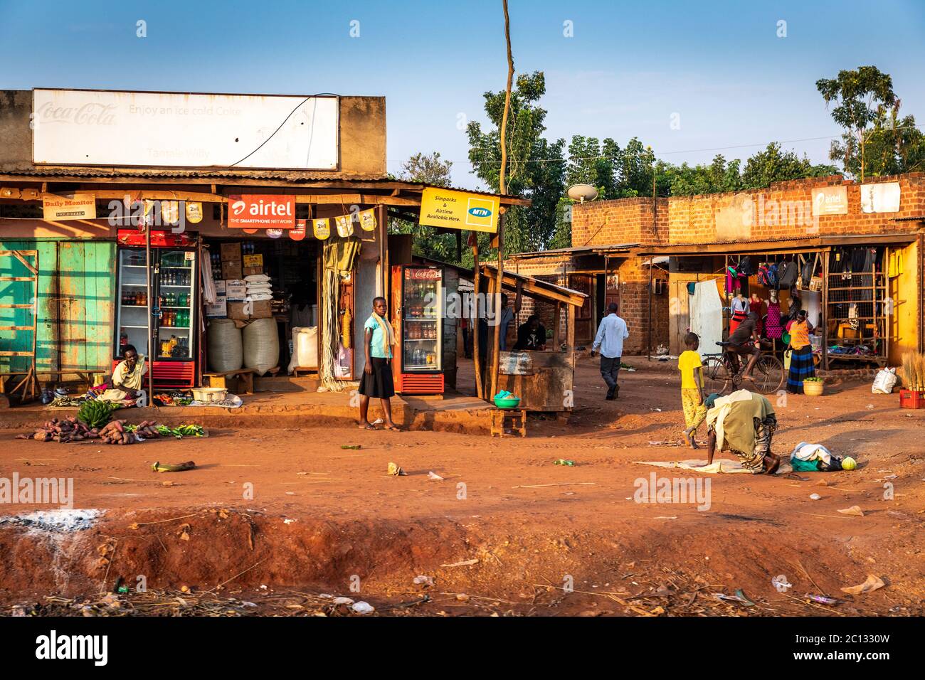 Roadside shops in rural Ugandan village with children in the street and a red dirt road, Uganda, Africa Stock Photo