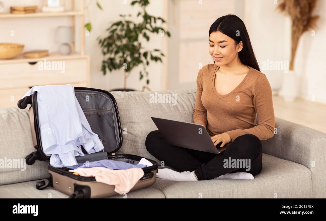 Chinese Girl Using Laptop Sitting With Unpacked Suitcase At Home Stock Photo