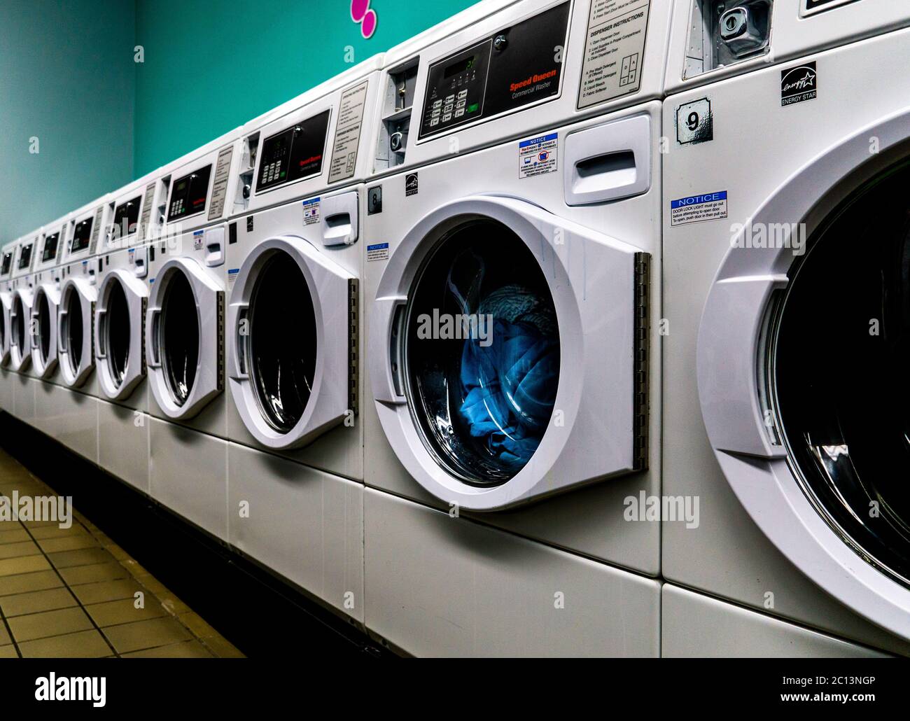 Laundromat line of white washing machines with laundry in them and is empty with teal wall and tile floor. Stock Photo