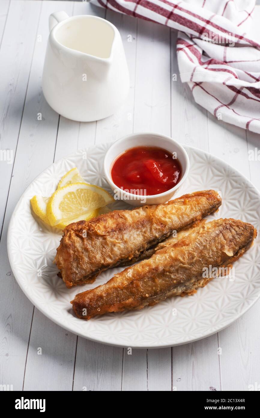 Fried fish hake Pollock with tomato sauce and slices of fresh lemon. Concept eating fatty fast food. White wooden table with Copy space Stock Photo