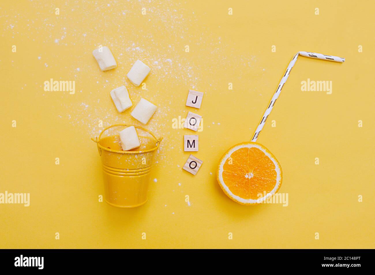 Abbreviation word JOMO written on of wood eco-friendly letters and festive cocktail tube in juicy ripe orange and marshmallows from a small yellow buc Stock Photo