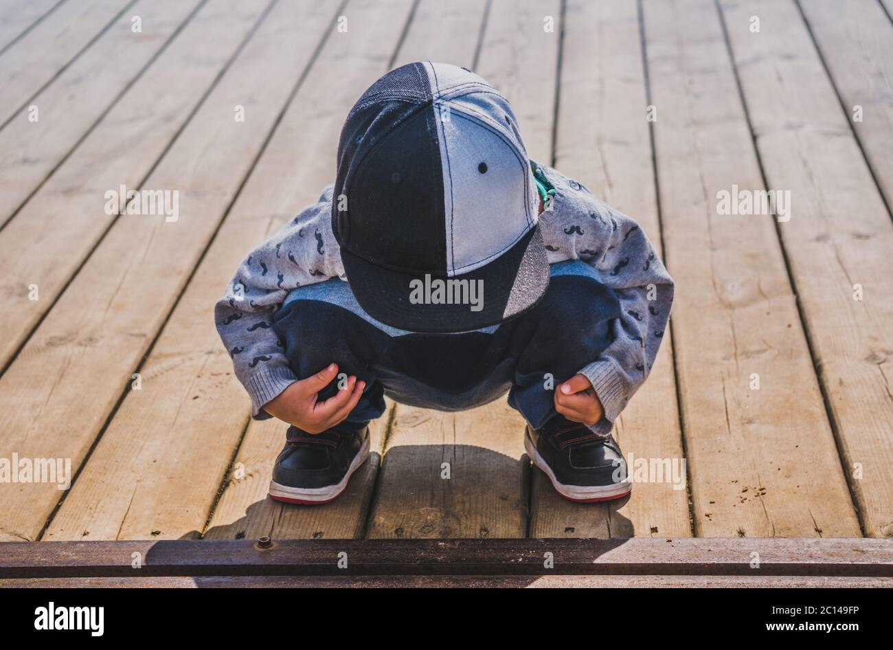 Small boy in baseball cap sitting in squatting position on wooden planked surface Stock Photo