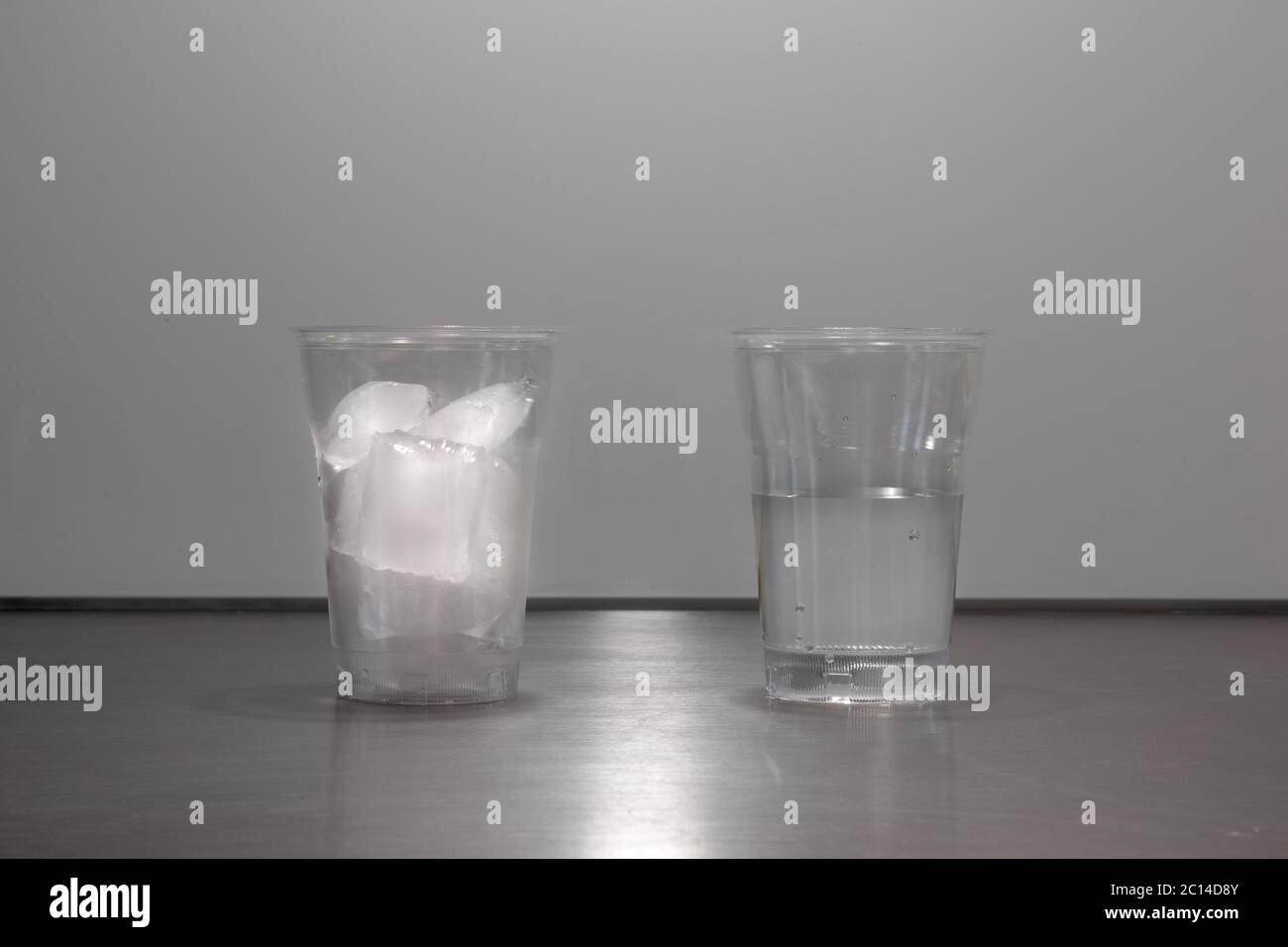 Ice cubes melting in glass or plastic cup showing change in physical state of water from ice to liquid. One cup with ice. One cup with water. Stock Photo