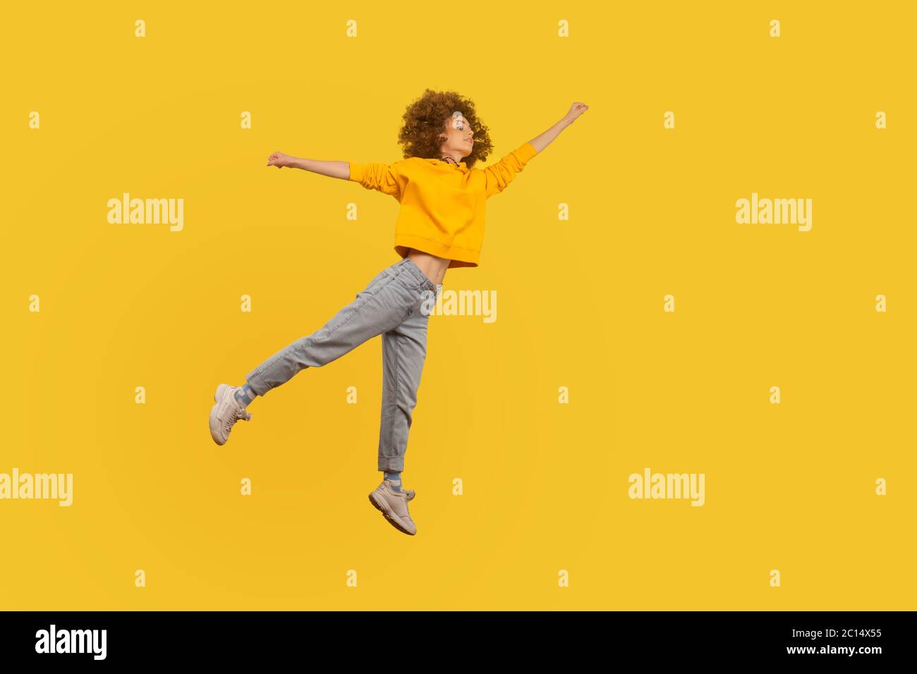 Portrait of superhero curly-haired girl in urban style outfit flying with raised hand high in air, feeling superpower and inspiration, achieving goal. Stock Photo