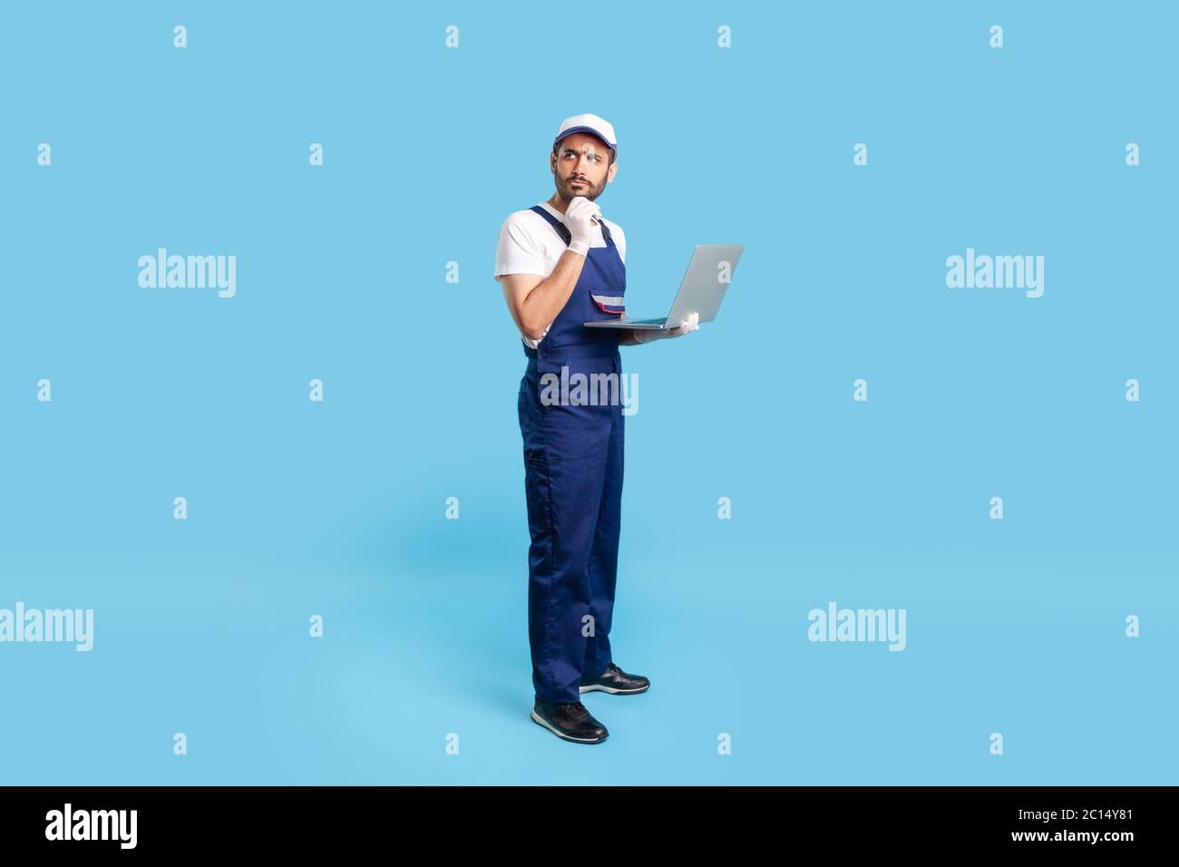 Full length thoughtful uncertain workman in uniform and protective gloves holding laptop, pondering decision with doubtful expression, online order fo Stock Photo