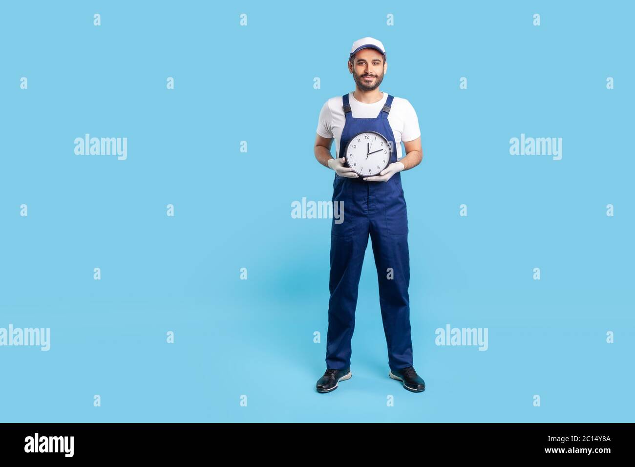 Full length optimistic punctual labourer in uniform holding clock and smiling to camera. Advertisement of professional house repair services on time. Stock Photo