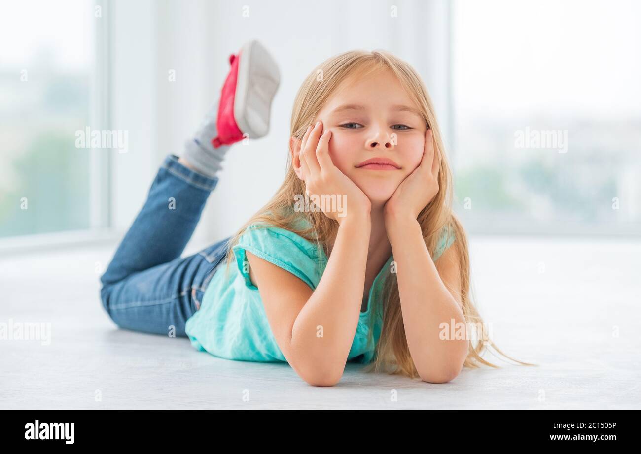 A blonde little girl in blue dress and fancy jeans lying on the floor on her stomach, relaxing Stock Photo
