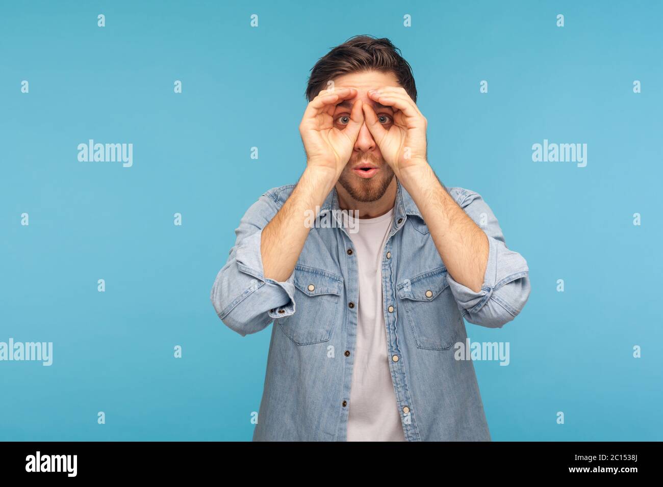 Wow, unbelievable discovery! Portrait of man in worker denim shirt looking far away through binoculars gesture, spying with shocked amazed expression. Stock Photo