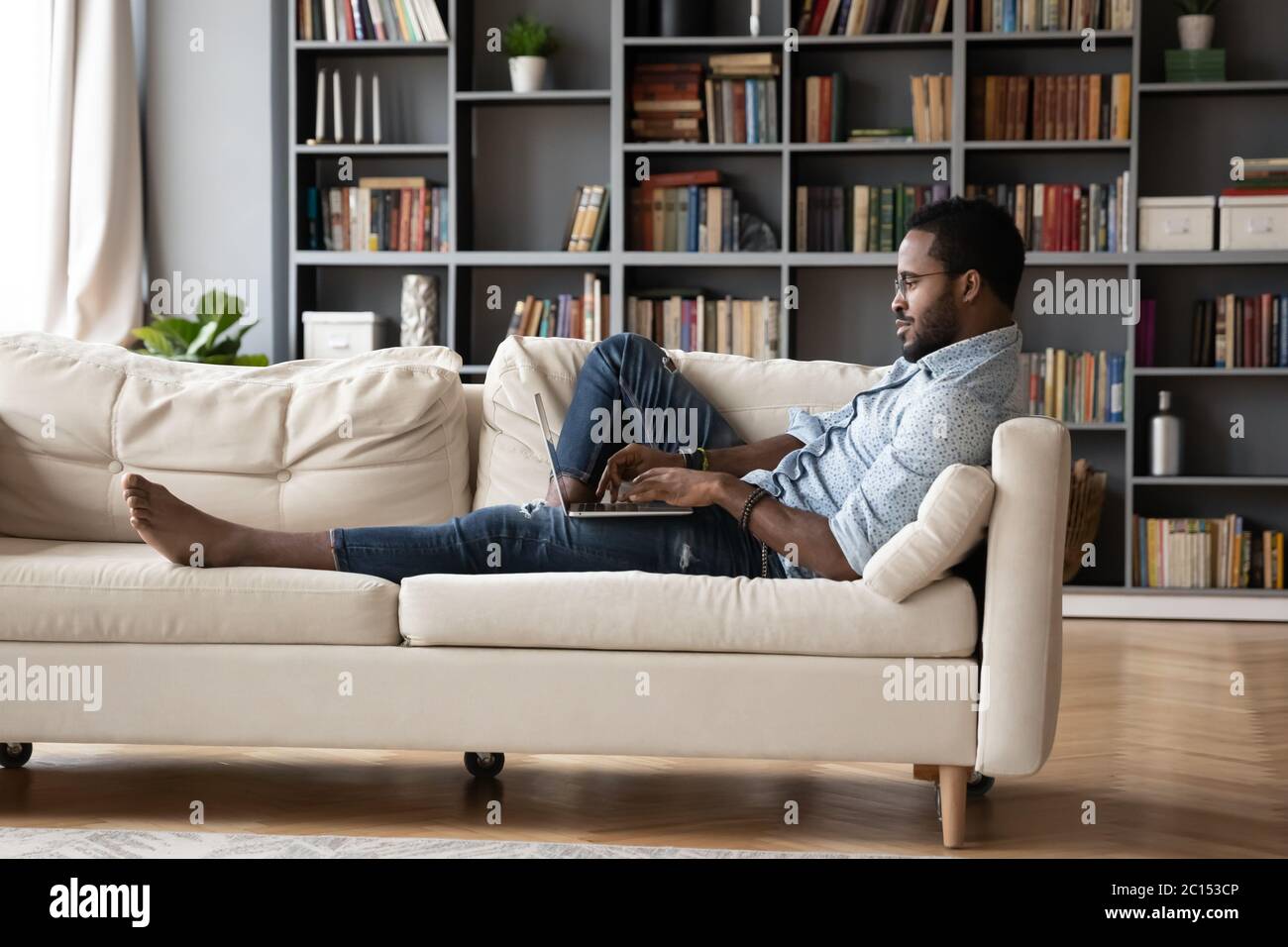 African guy lying on sofa typing message using computer Stock Photo