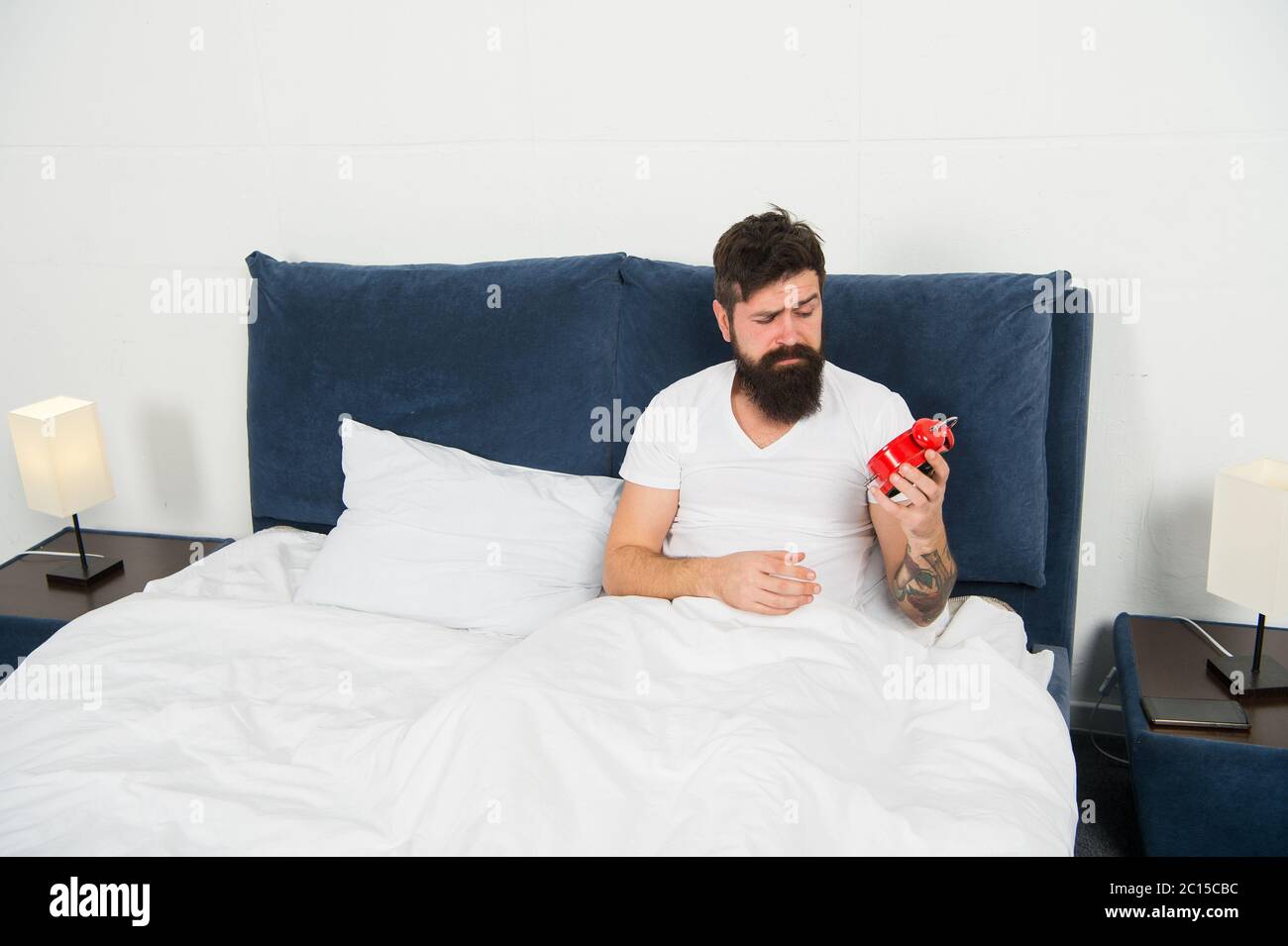 Its hard to get out of bed. Bearded man check time with alarm clock. Sleep time. Sleep and wake routine. Bed time. Bedtime. Morning and night. Regular time to go to bed and wake up. Stock Photo