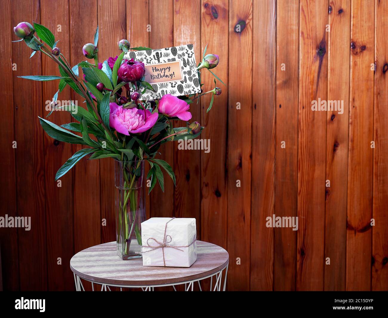 Pink flowers peonies in a vase on a wooden background with a small white gift and a happy birthday card. The photo was taken with natural daylight. Stock Photo