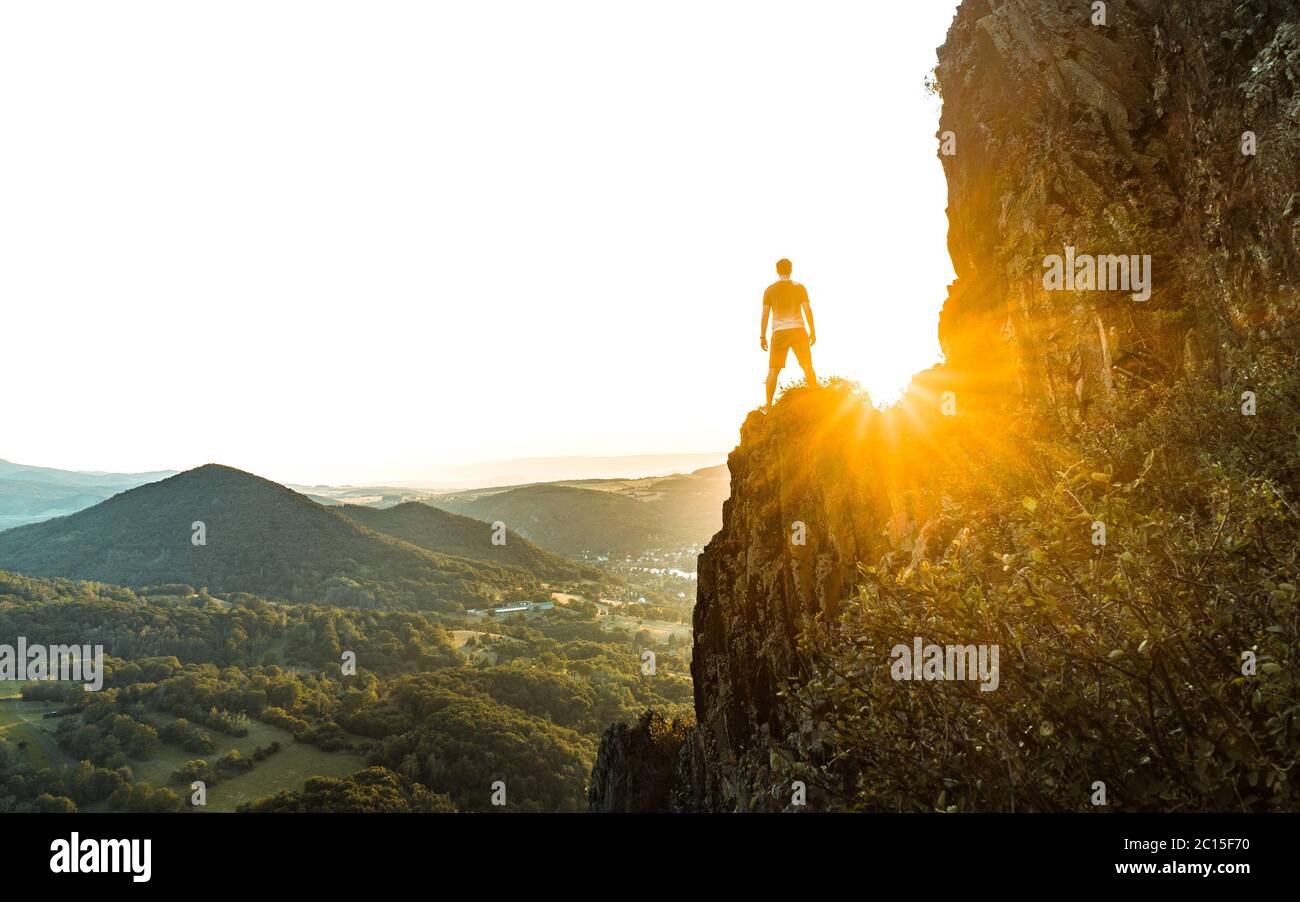 Silhouette of the person on the high rock at sunset. Satisfy hiker enjoy view. Tall man on rocky cliff watching down to landscape. Vivid and strong Stock Photo