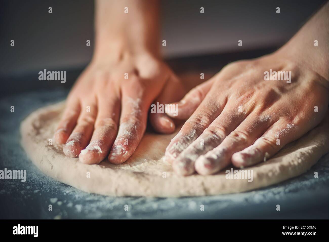 A man with his hands rolls out homemade pizza dough, lying on a dark baking tray and illuminated by light. Cooking at home. Stock Photo