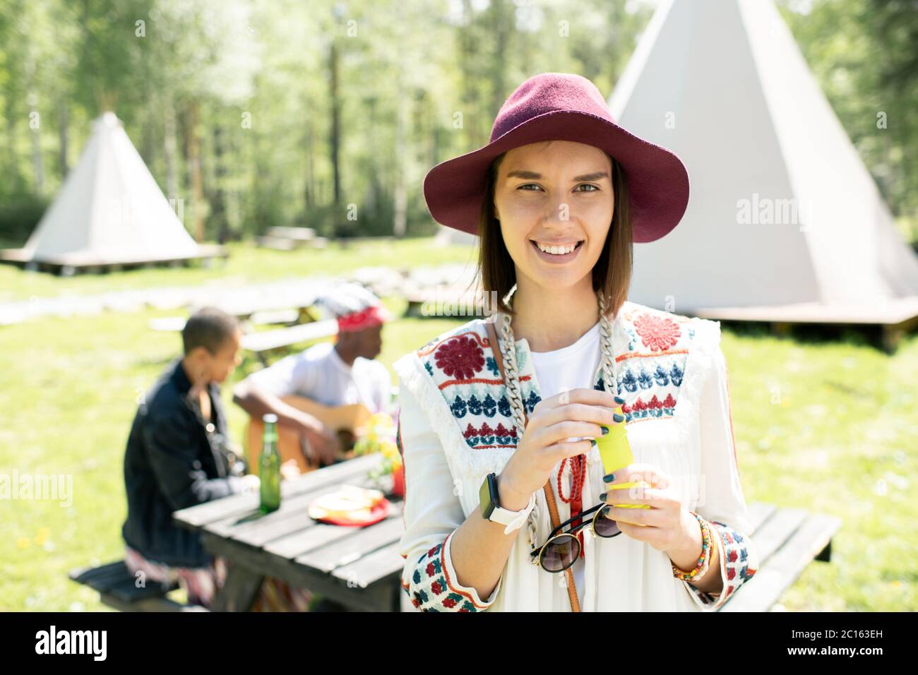 Portrait of happy attractive hippie girl in beautiful hat standing against friends table at festival campsite Stock Photo
