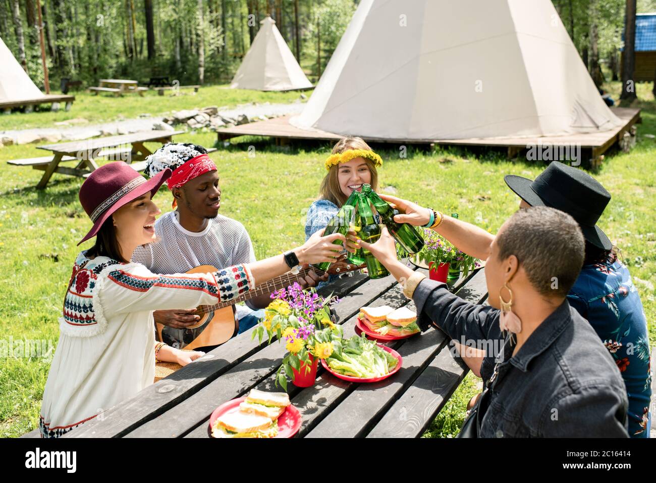Group of positive multi-ethnic friends sitting at table with snacks and drinking beer together at campsite Stock Photo