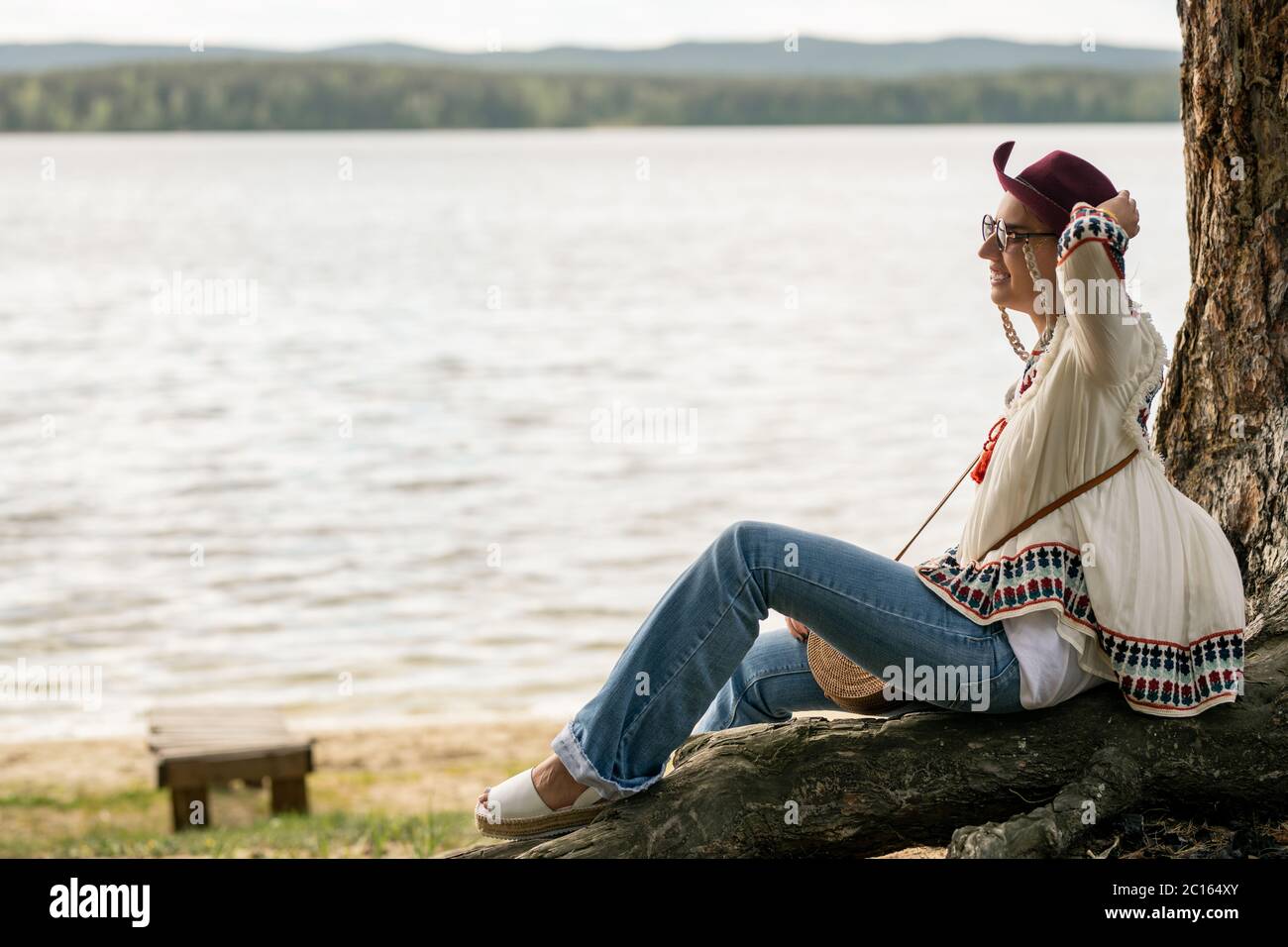 Smiling young woman in eyeglasses and hippie blouse sitting at tree and holding hat while enjoying wind near lake Stock Photo