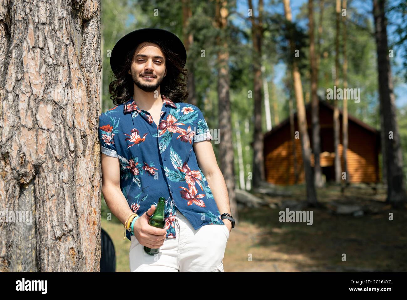 Portrait of positive hippie guy in flower shirt and hat leaning on tree and holding beer bottle in forest Stock Photo
