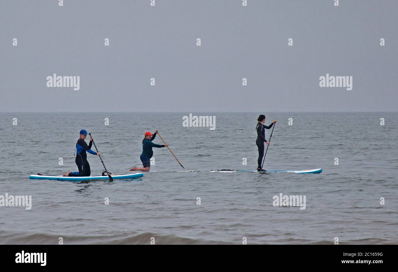 Portobello, Scotland, UK. 14 June 2020. After yesterday's fog all day, it turned a little brighter  and warmer today but still dreich with cloudy skies, temperature of 16 degrees centigrade. Busy seaside with all the cafes and pub open, pictured: three female paddle boarders and one family at the seashore. Stock Photo