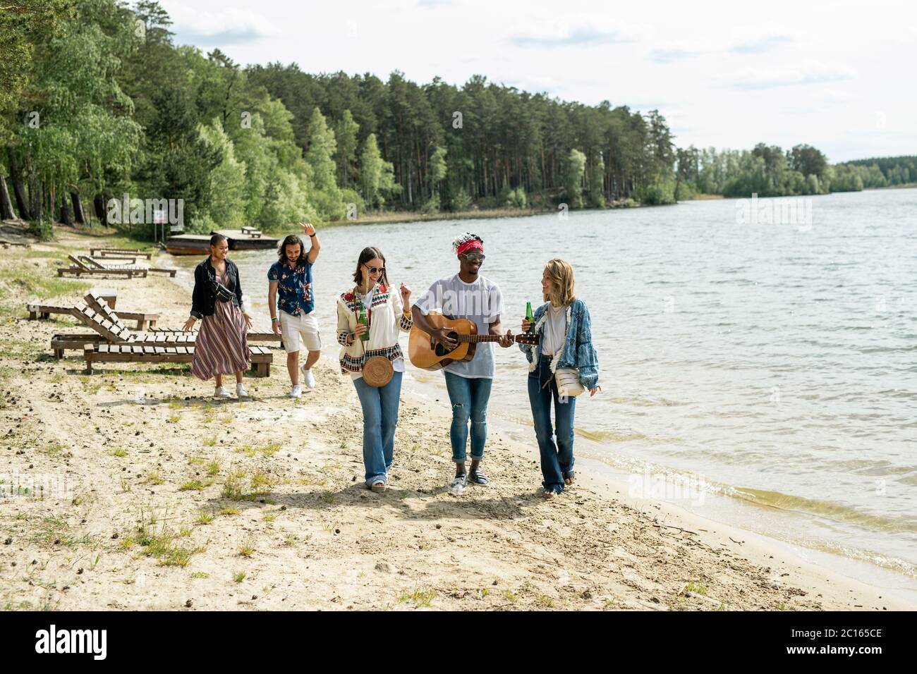 Group of positive young multi-ethnic friends enjoying beach stroll while black guy playing guitar on move Stock Photo