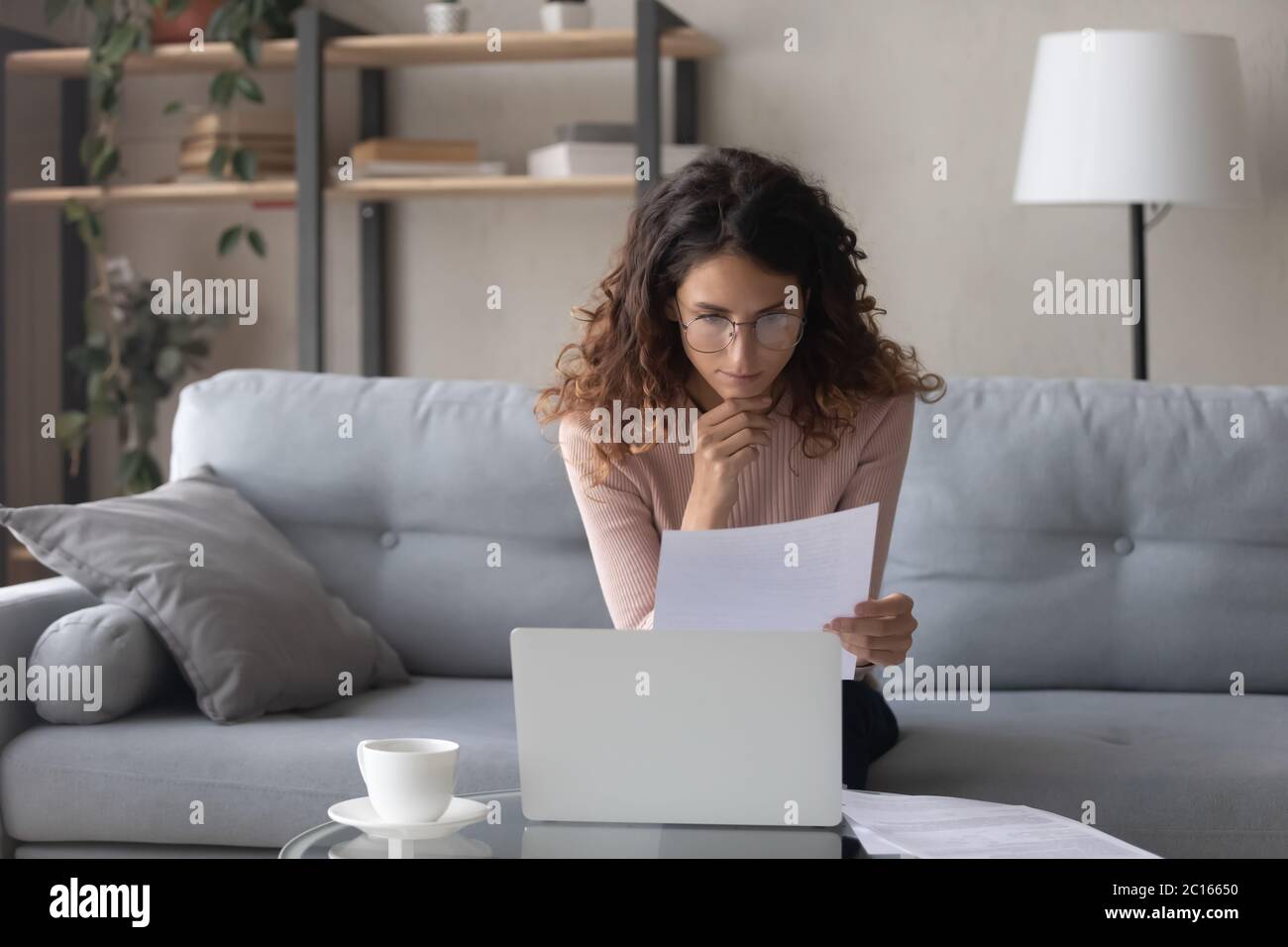 Thoughtful woman wearing glasses reading document, using laptop Stock Photo