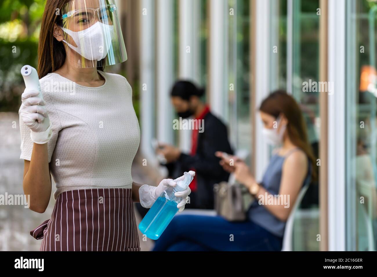 Portrait attractive asian waitress wear face mask and shield hold thermometer and alcohol gel sanitizer with background of social distancing customer Stock Photo