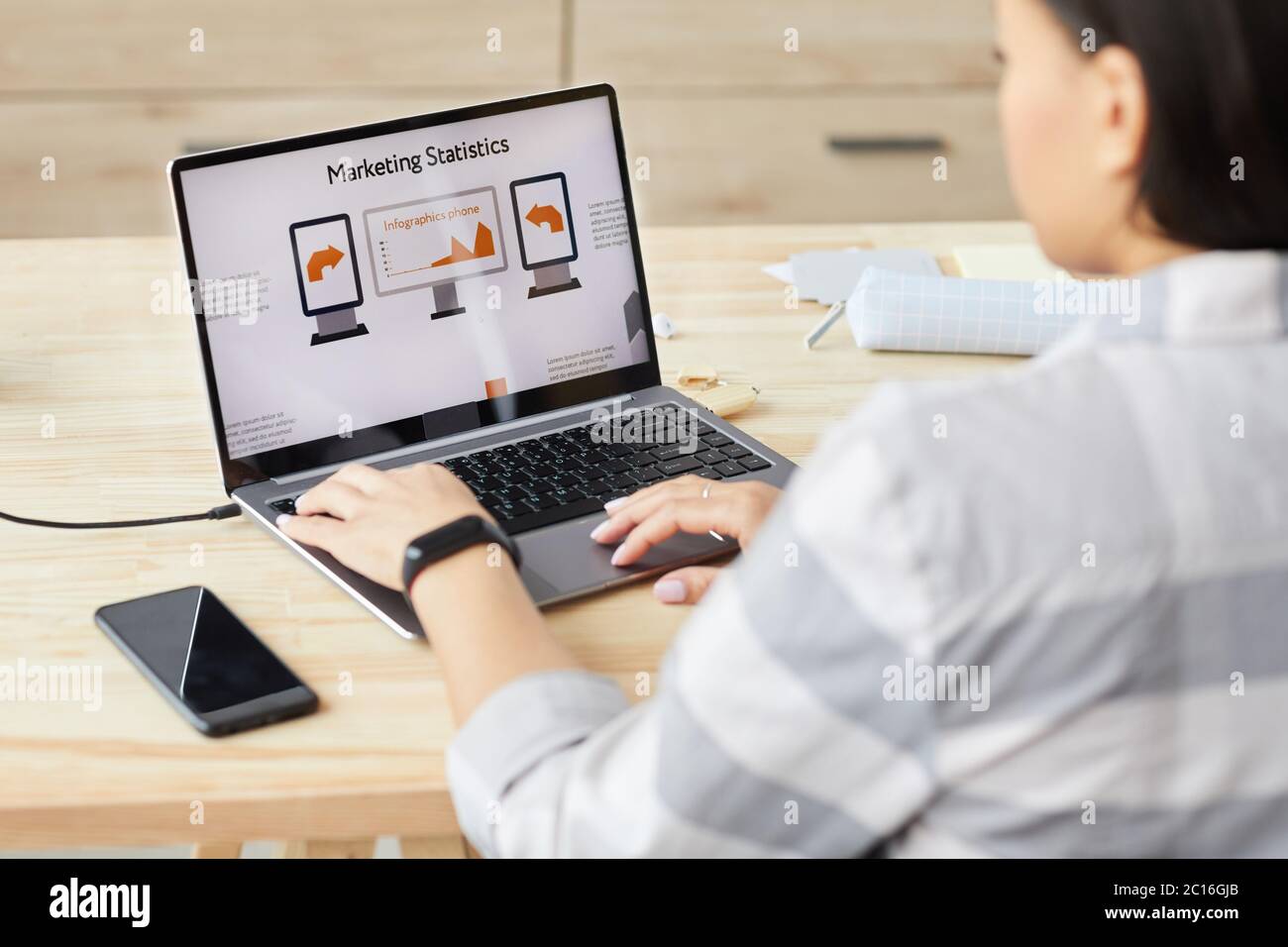 High angle view at young woman looking at laptop screen with Marketing Statistics headline while sitting by wooden table and working at home office, c Stock Photo