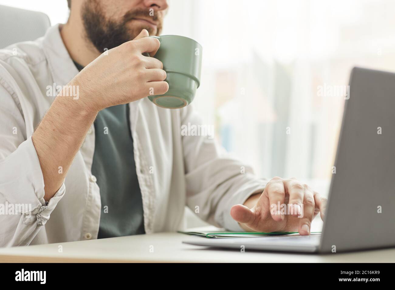 Cropped portrait of bearded adult man drinking coffee while using laptop at home office, copy space Stock Photo