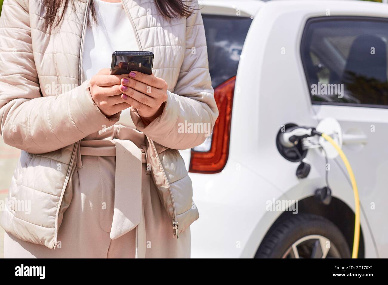 Woman with a mobile phone near recharging electric car. Vehicle